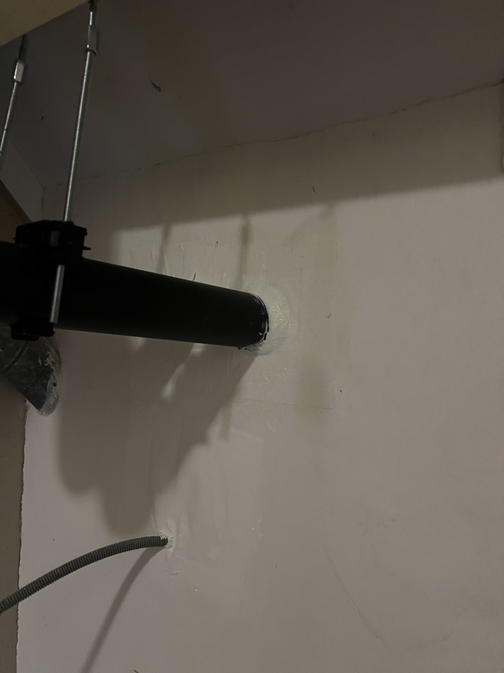 Een zwarte buis komt door een wand naar binnen, met daarnaast een grijze flexibele slang en metalen ophangbeugels.