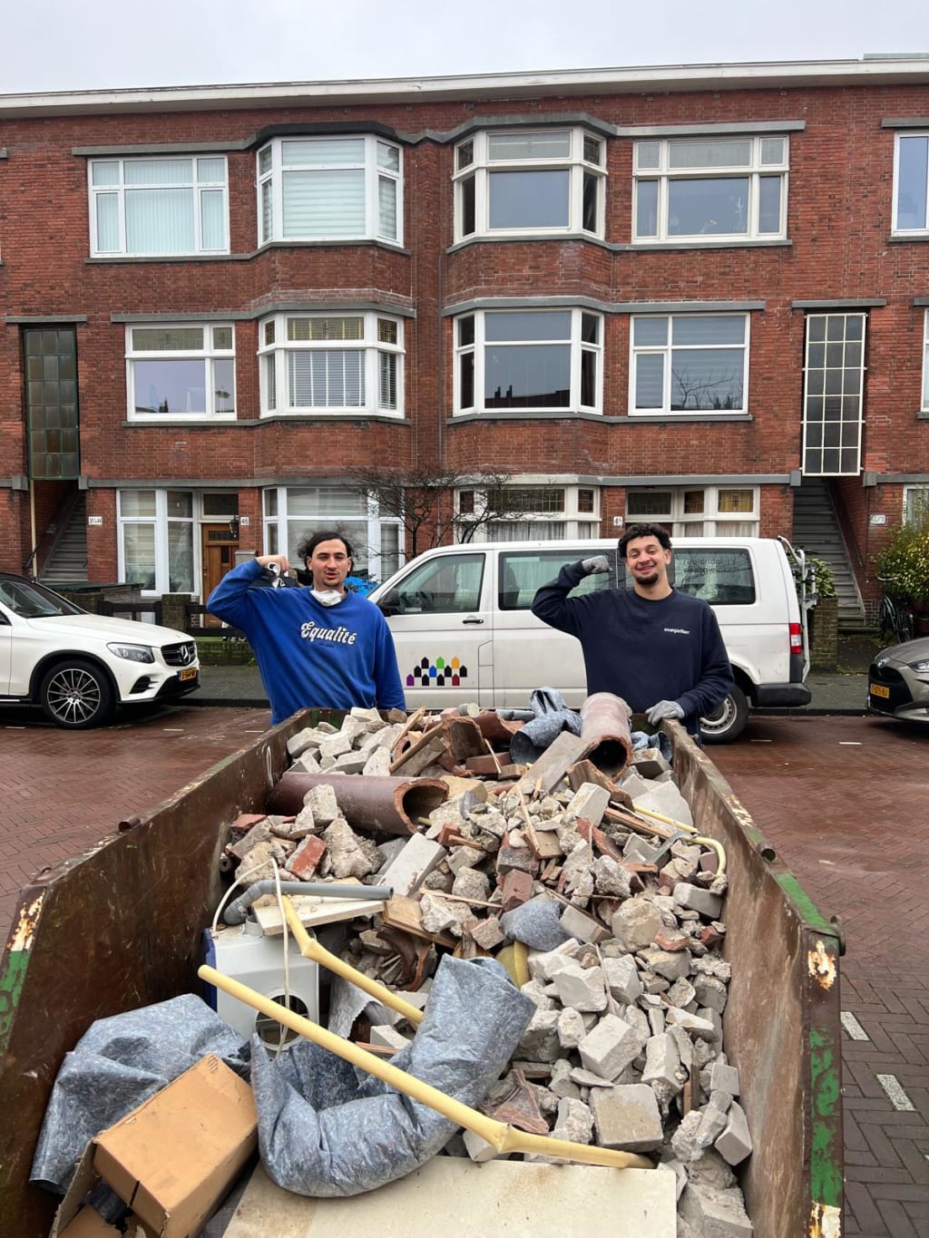 Twee mannen staan glimlachend achter een volle container met bouwpuin op een straat met bakstenen huizen.