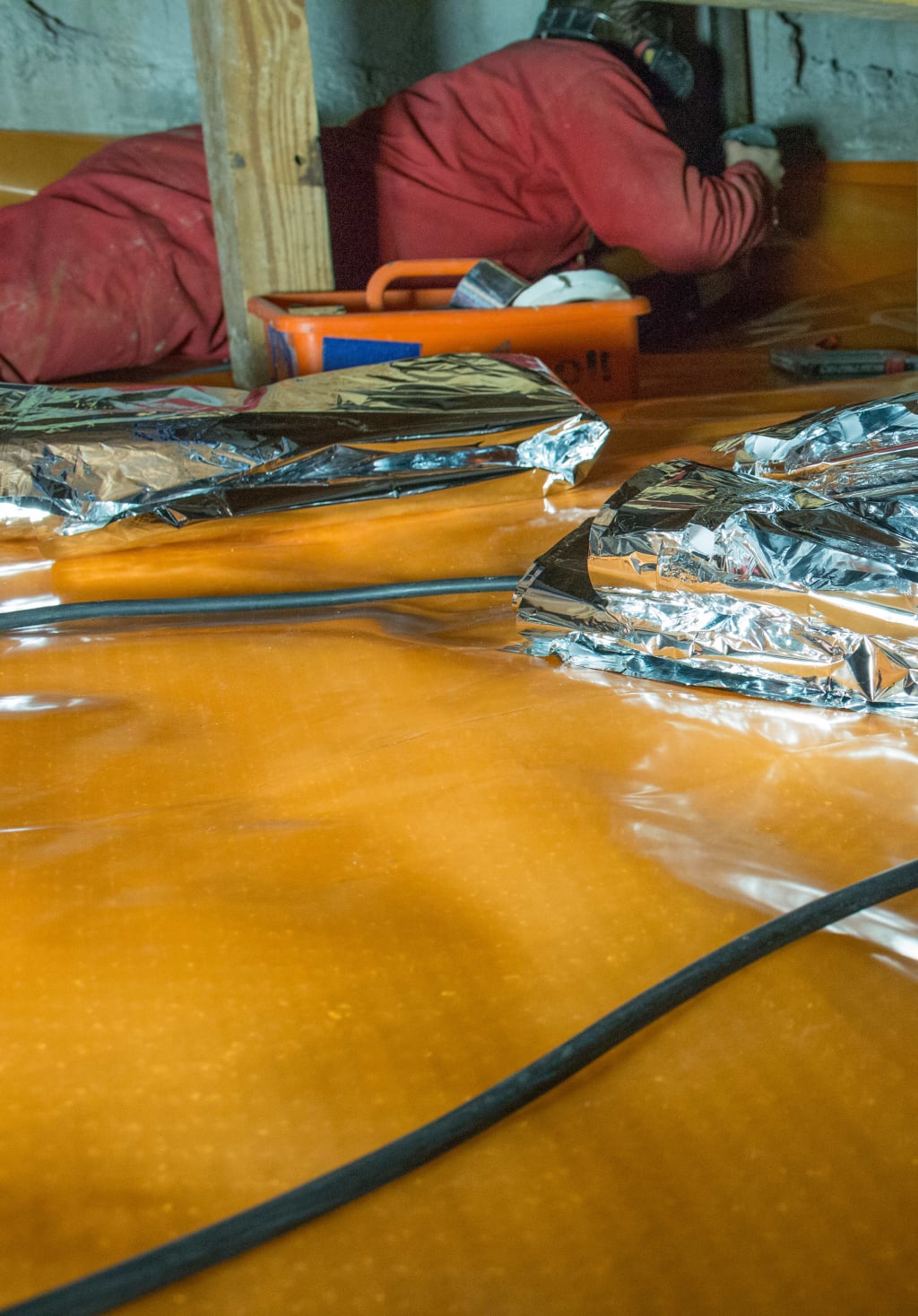 Een persoon in een rode overall werkt in een kruipruimte met oranje folie en zilverkleurige isolatiemateriaal op de vloer.