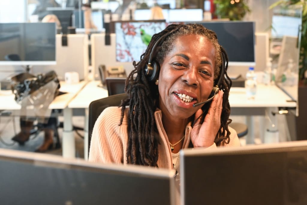 Een persoon met een glimlach die aan het telefoneren is met een hoofdtelefoon op. Op de achtergrond zijn computers te zien.