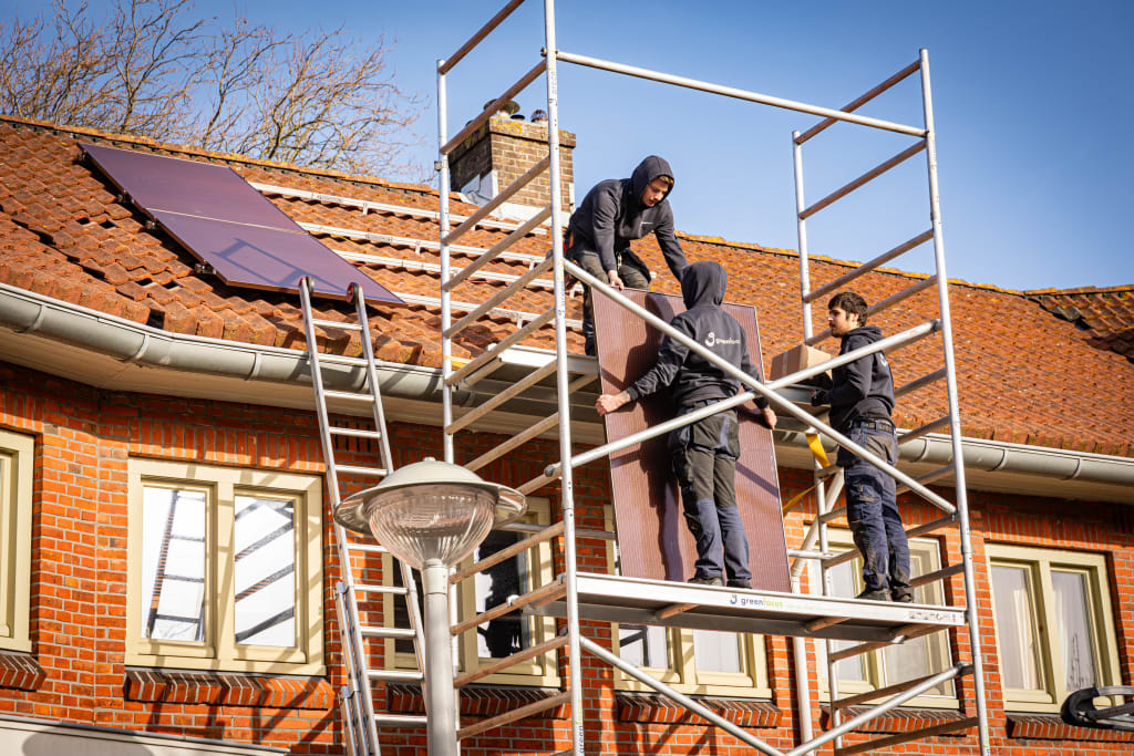 3 personen op een stijger bij een woning die zonnepanelen aan het plaatsen zijn op het dak van het huis. Twee op de stijger die spullen aangeven aan iemand op het dak.