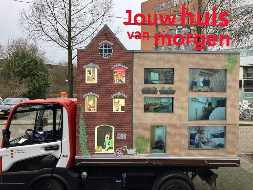 Een super klein busje waar de achterkant een bak heeft waar een knutselwerk van Jouw huis van morgen op staat. Daar zie je een woning met in de ramen afbeeldingen van woningen.