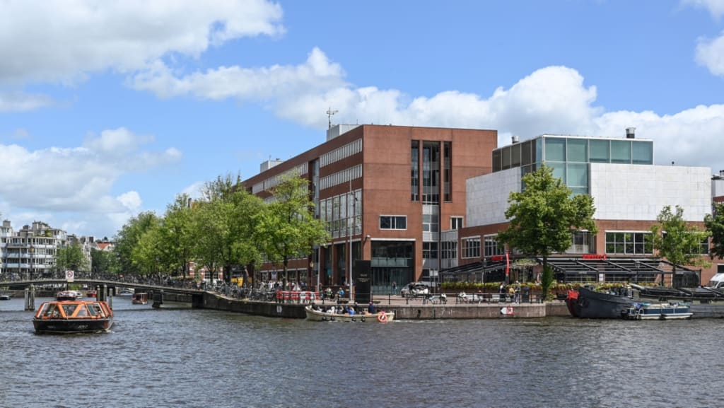 Amsterdamse grachten met verschillende bootjes die varen. Dan de kade met een groot gebouw en groene bomen ervoor.