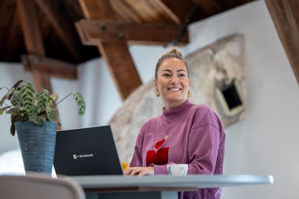 Een persoon die achter een bureau met een laptop erop staat. Diegene lacht ergens naar. Op het bureau staat een plant.