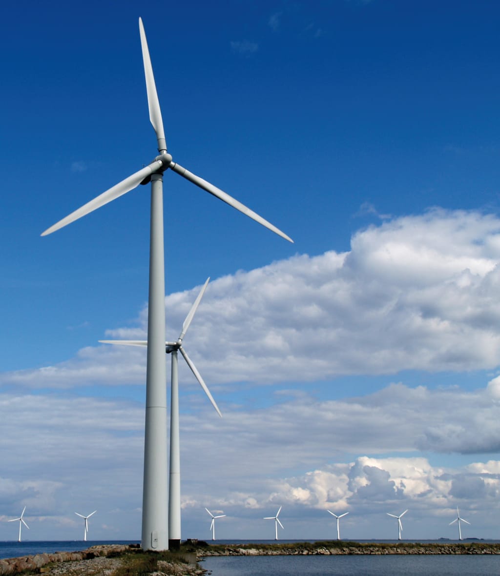 Een rij windmolens. Een grote op de voorgrond en kleinere op de achtergrond. De windmolens staan aan water.