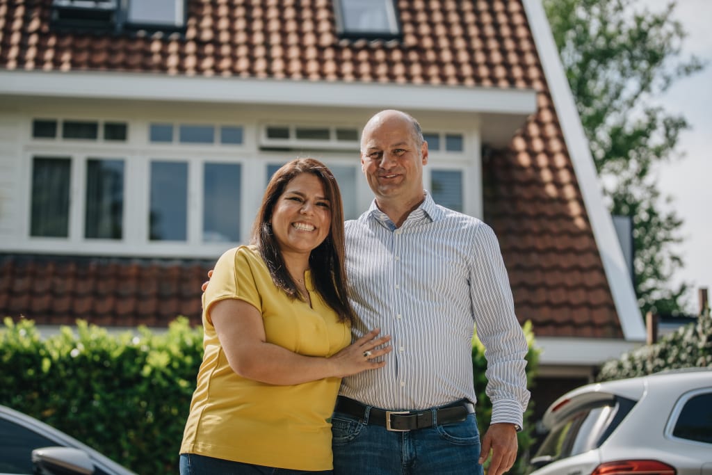 Foto van een vrouw van kleur en een witte man die samen tussen twee auto's voor een huis staan. Ze lachen naar de camera.