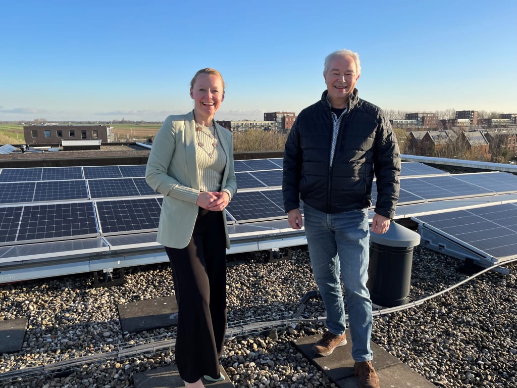 Twee mensen die op een dak staan. Achter hen zijn een heleboel zonnepanelen te zien. Ze lachen beiden naar de camera.
