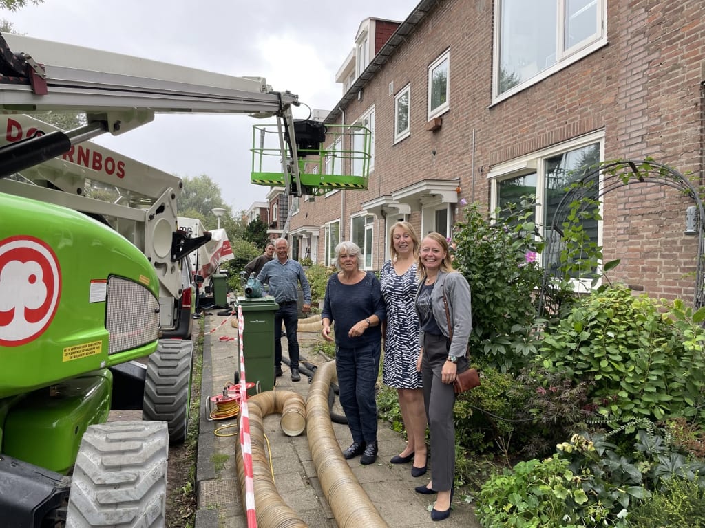 Een groep mensen staat op de stoep voor hun woningen waar de stoep is afgezet en constructie machines staan.