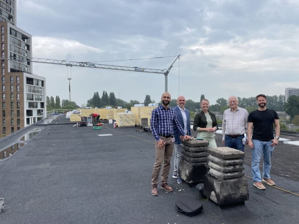 Een groep van 5 mensen staat op een plat dak met tegels op de voorgrond en constructie op de achtergrond