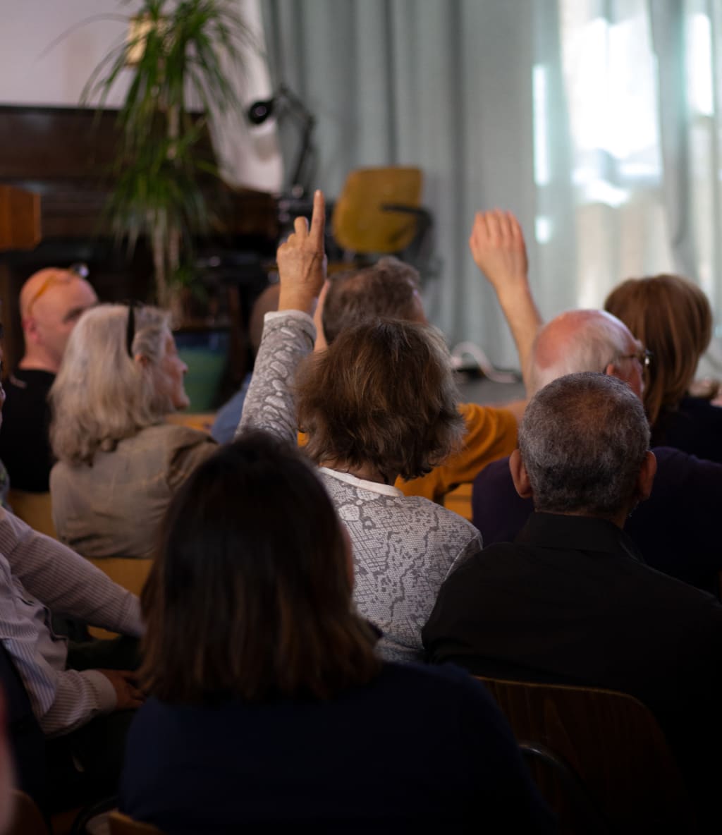 Een groep aanwezigen in een zaal, waarvan sommigen hun hand opsteken tijdens een interactieve sessie.