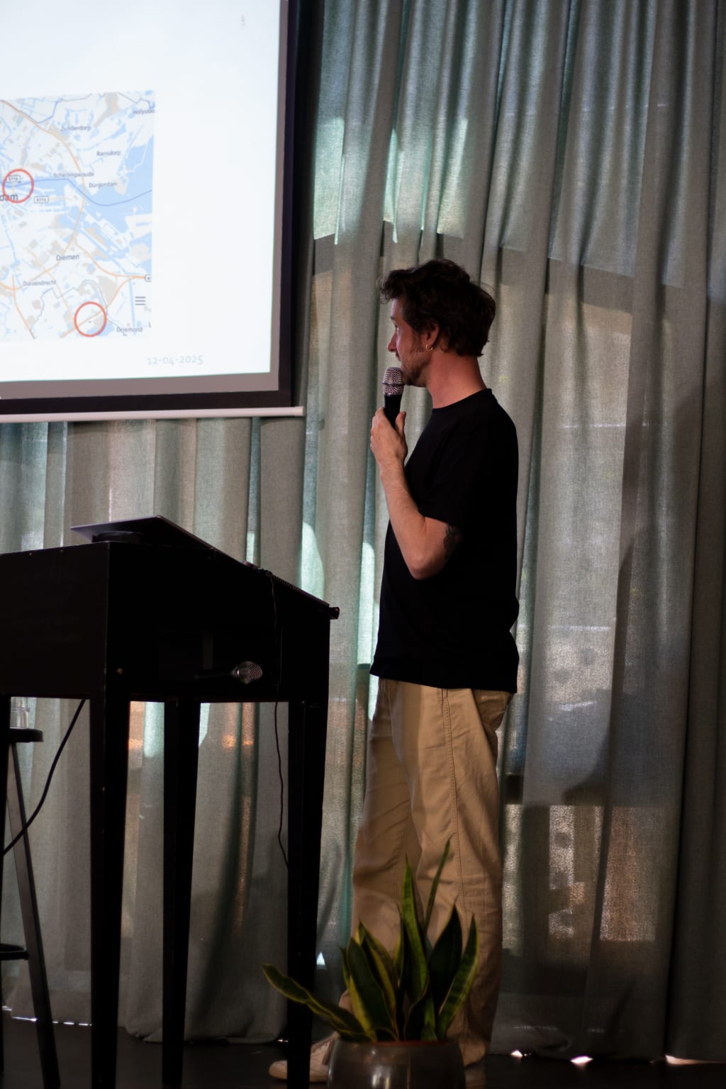 Een spreker met microfoon geeft een presentatie over verduurzaming, terwijl een kaart van Amsterdam met gemarkeerde locaties op het scherm wordt getoond.