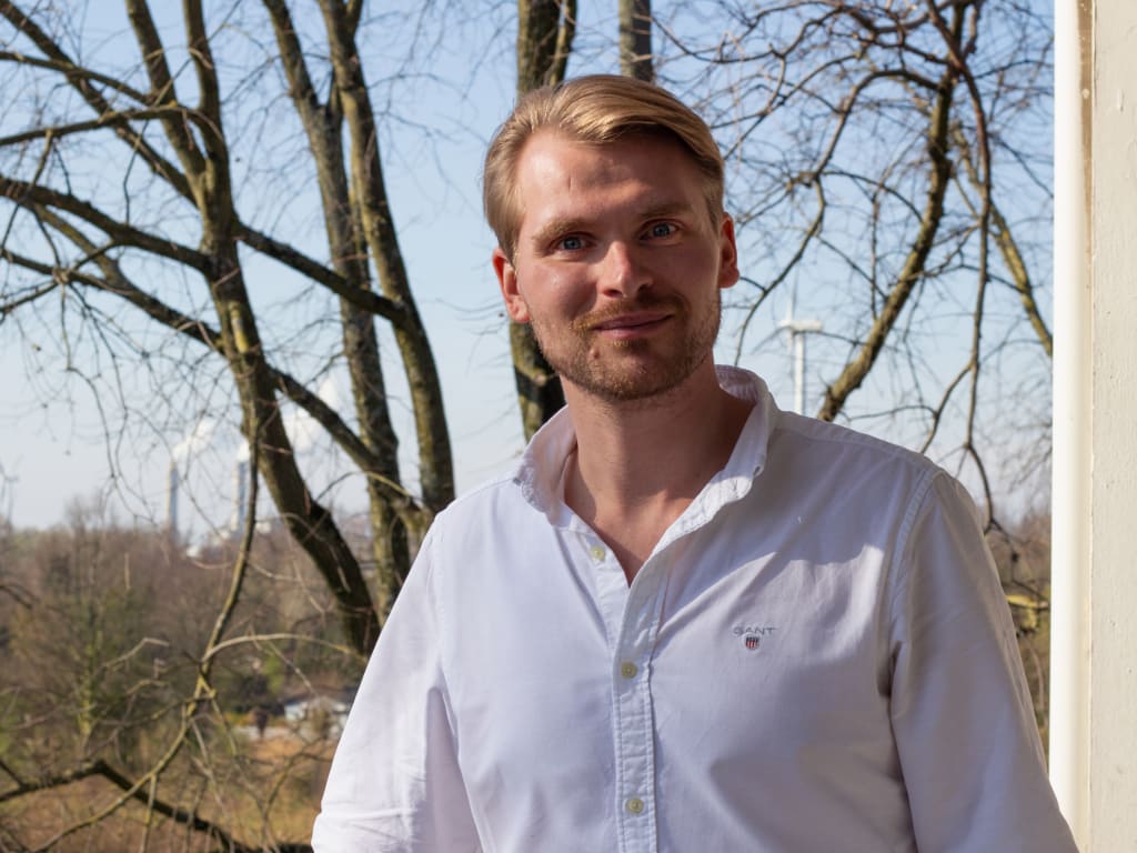 Een man in een wit overhemd staat op een balkon met bomen en windturbines op de achtergrond.