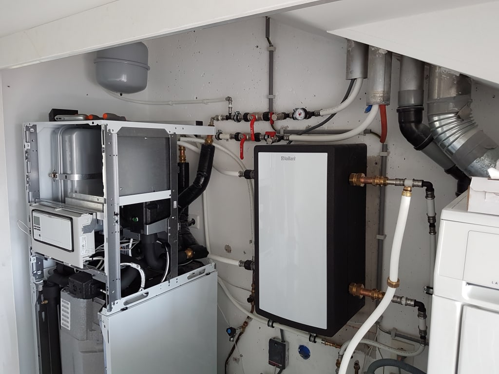 Een verwarmingssysteem in een technische ruimte, met een Vaillant-warmtepomp, leidingen en een waterdrukmeter.