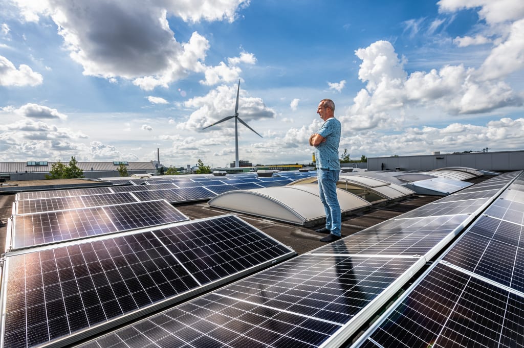 Man staat op een dak met zonnepanelen terwijl een windturbine op de achtergrond draait, onder een deels bewolkte blauwe lucht.