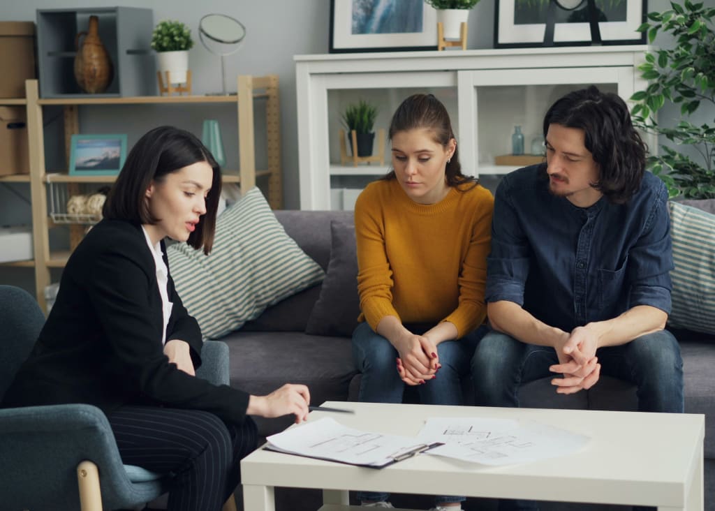 Een vrouw in pak legt papieren uit aan een jong stel dat aandachtig luistert op een bank in een huiskamer.