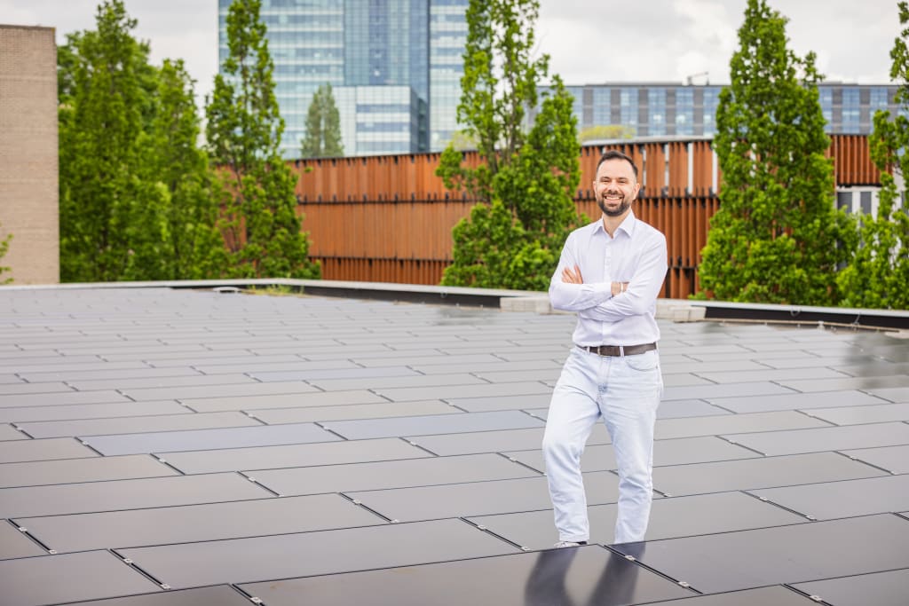 Glimlachende man in witte blouse staat op een dak met zwarte zonnepanelen, met moderne gebouwen en bomen op de achtergrond.