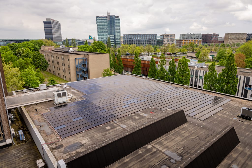 Luchtfoto van een groot plat dak bedekt met zonnepanelen, omringd door stedelijke bebouwing en groene bomen.