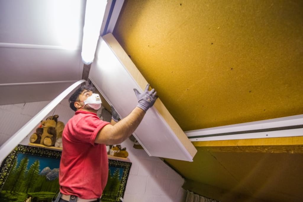 Een man met een mondkap op is bezig met het plaatsen van isolatie tegen de gele schuine muur van de zolder