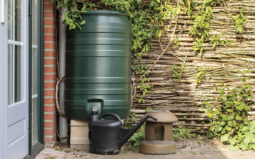 Een foto van een groene regenton naast een huis, met daarbij staand een zwarte gieter.