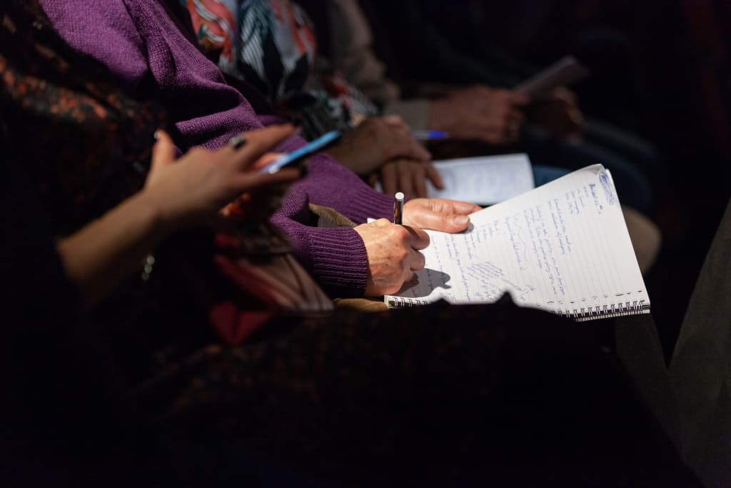 Een hand schrijft in een notitieboek terwijl andere mensen hun telefoons vasthouden, met een focus op het notitieboek en de hand die schrijft.
