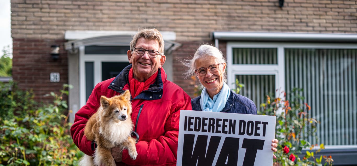 Twee witte mensen die voor hun huis staan. Ze houden hun hond en een bord met 'iedereen doet wat' erop vast.