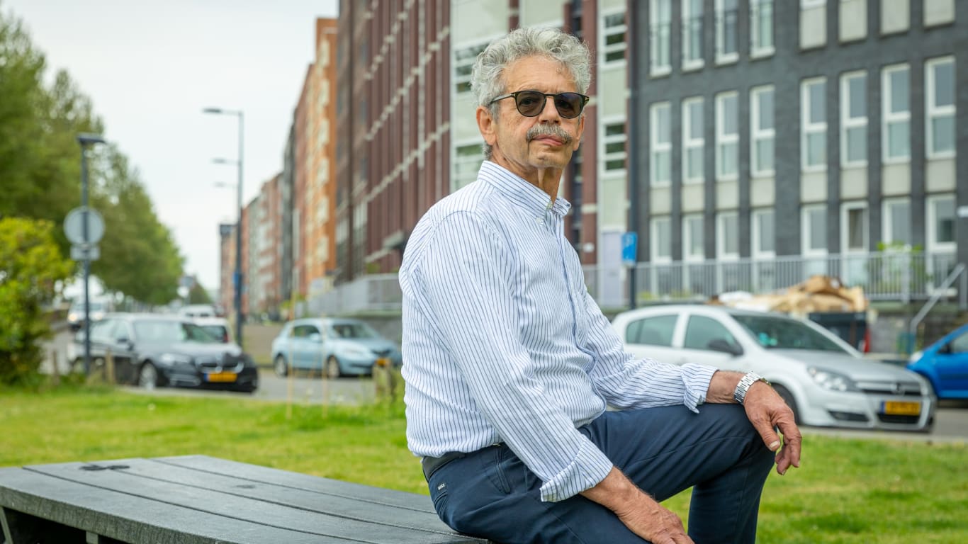 Een man die op een bank zit met een stadsachtergrond en auto's op de achtergrond, terwijl hij naar de camera kijkt.
