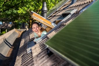 Een witte vrouw die vanuit haar dakraam haar hoofd uitsteekt en naar de zwarte zonnepanelen op het dak kijkt.