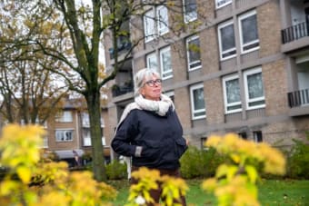 Een witte vrouw met zwarte jas en witte sjaal loopt voor een flatgebouw, buiten. Op de voorgrond zijn gele bloemetjes te zien.