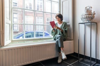 Een witte vrouw die in haar raamkozijn zit. Onder het raamkozijn zit een verwarming en de vrouw kijkt naar een tablet.