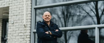 Een foto van een witte man met een zwarte jas en broek die voor zijn huis staat met de handen over elkaar. Hij lacht naar de camera.