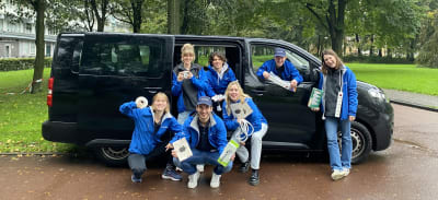 Een groepje mensen die in blauwe jasjes bij een zwart busje staan. Ze houden allemaal energiebesparende maatregelen vast en kijken en lachen in de camera.