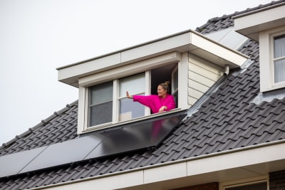 Een dak met zonnepanelen en een mevrouw zwaaiend uit haar dakraam.