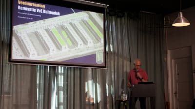 Een man geeft een presentatie over verduurzaming van een gebouw, met een architectonisch ontwerp op het scherm.