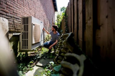 Man knielt buiten in een smalle ruimte tussen een bakstenen muur en houten schutting en werkt aan een grote airconditioningunit.