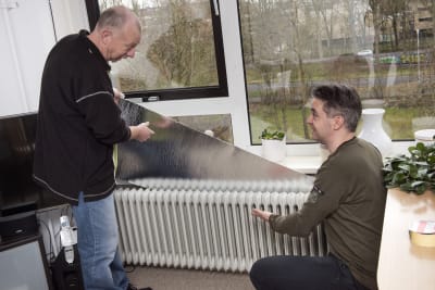 Twee witte mannen plaatsen radiatorfolie achter een witte radiator. Eén man staat en de andere is gehurkt.