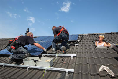 Twee vakspecialisten die zonnepanelen aan het leggen zijn en een mevrouw die uit haar dakraam hiernaar kijkt.