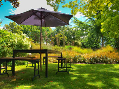 Parasol boven tafel met 4 bankjes op groen grasveld met bomen er omheen