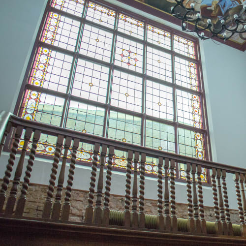 Groot glas-in-lood raam in karakteristiek oud pand met houten balustrade op de voorgrond en kroonluchter aan het plafond