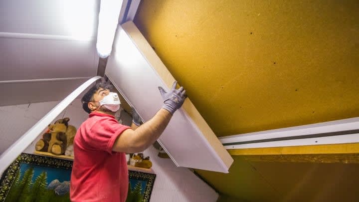 Een man met een mondkap op is bezig met het plaatsen van isolatie tegen de gele schuine muur van de zolder